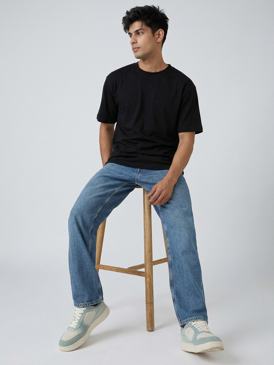 Man sitting on a wooden stool wearing a black t-shirt, blue jeans, and light-colored sneakers against a white background.