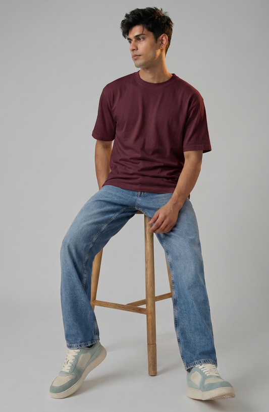 Man sitting on a wooden stool wearing a maroon t-shirt and blue jeans against a gray background