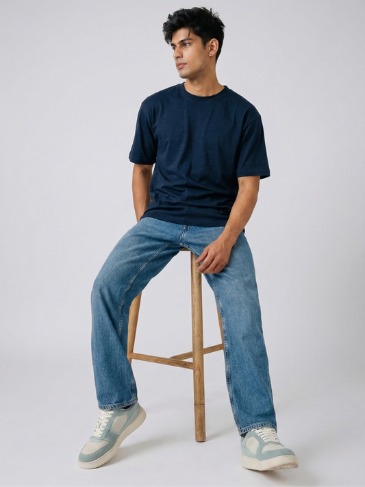 Man sitting on a wooden stool wearing a black t-shirt, blue jeans, and light-colored sneakers against a white background.