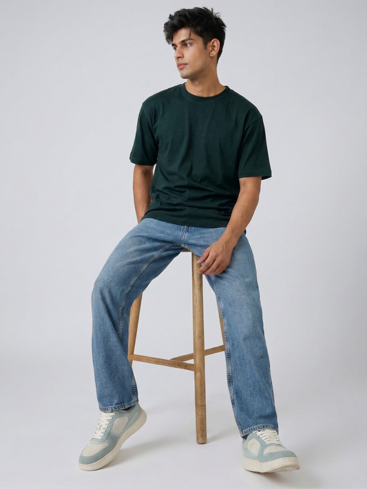 Man sitting on a wooden stool wearing a dark green t-shirt and blue jeans against a white background