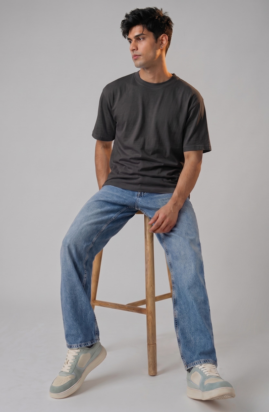 Man sitting on a wooden stool wearing a dark t-shirt and blue jeans against a gray background