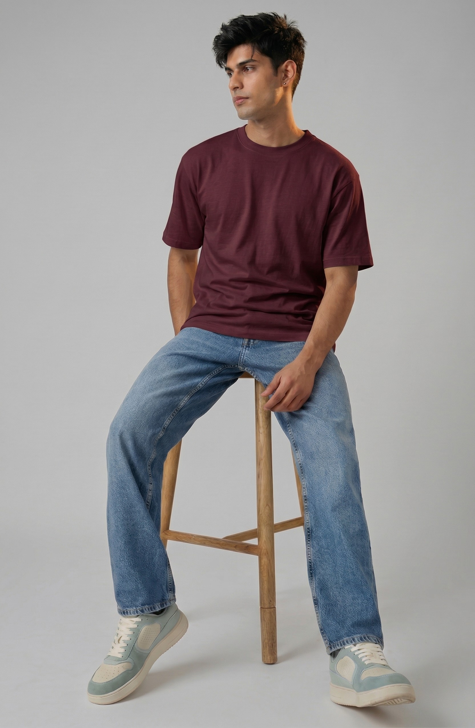 Man sitting on a wooden stool wearing a maroon t-shirt and blue jeans against a gray background