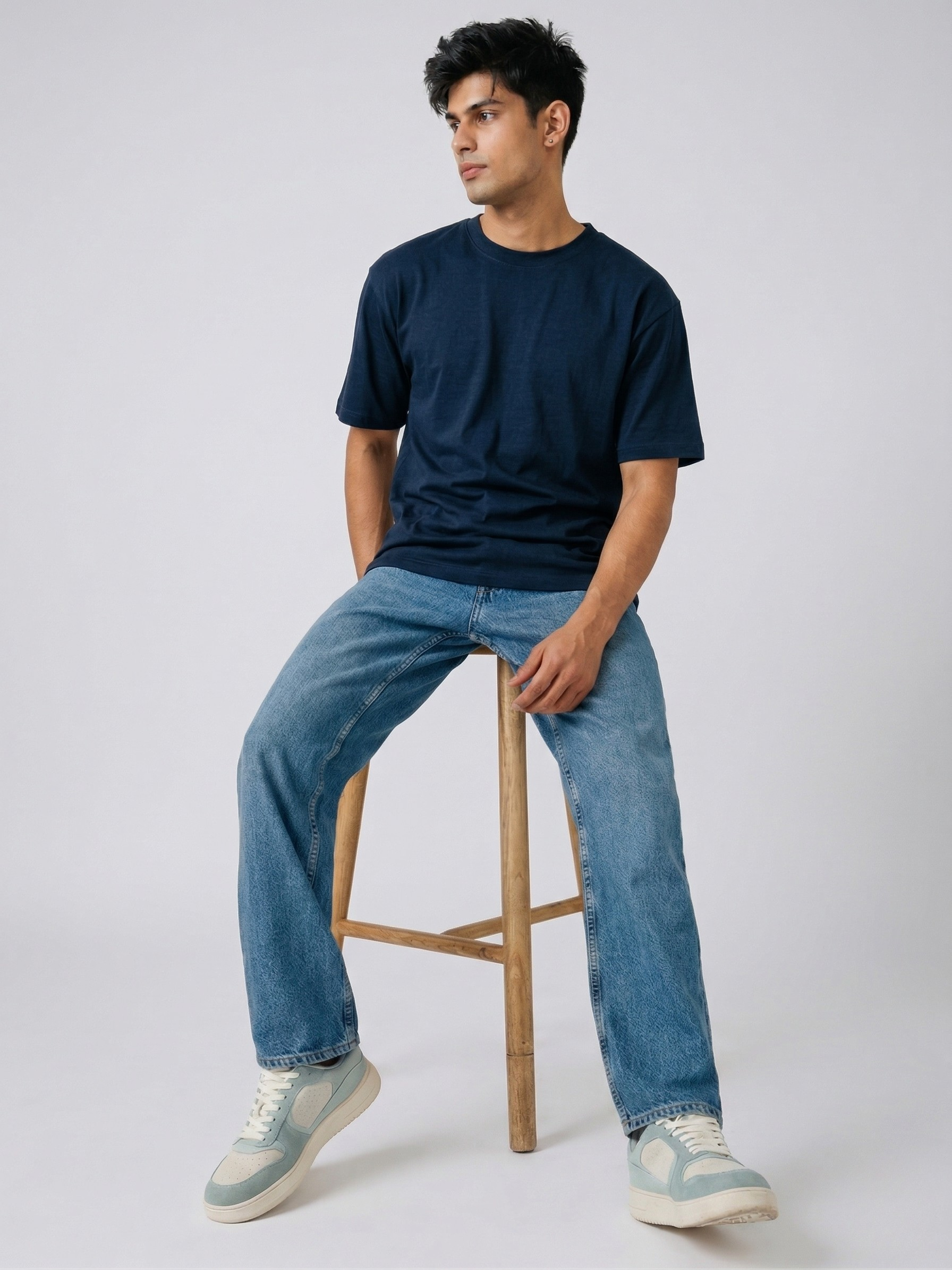 Man sitting on a wooden stool wearing a black t-shirt, blue jeans, and light-colored sneakers against a white background.