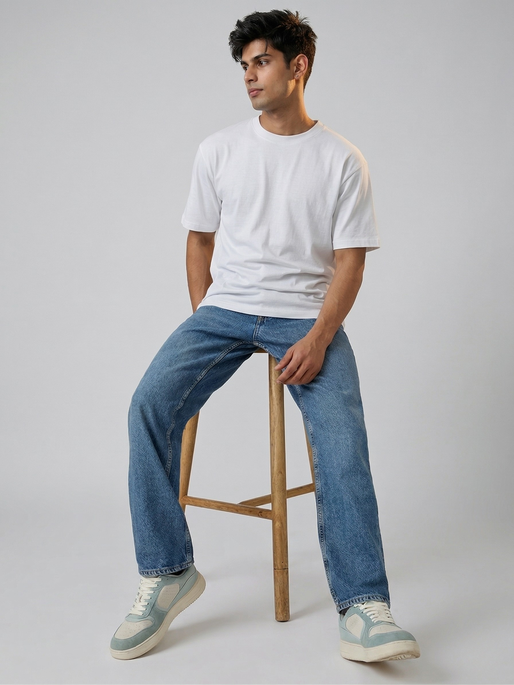 Man sitting on a stool wearing a white t-shirt and blue jeans against a gray background